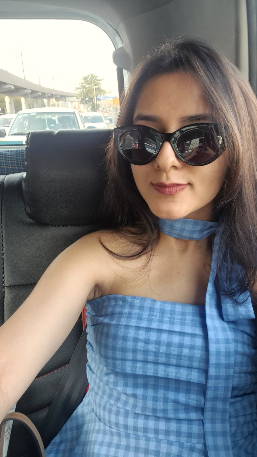 Labdhi Kothari in a blue gingham dress and black cat-eye sunglasses, taking a selfie in a car
