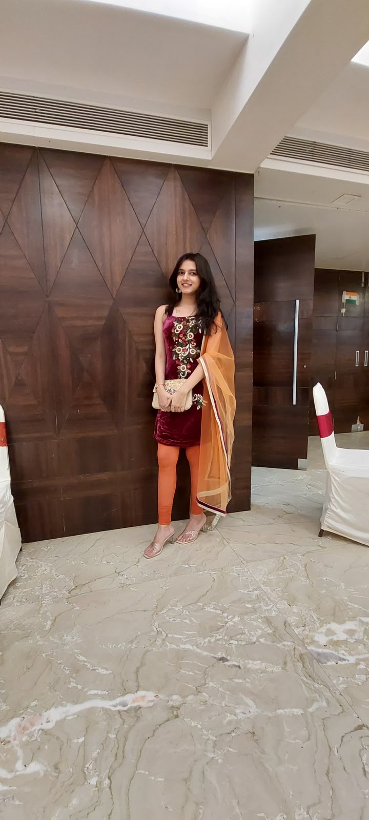 Labdhi Kothari in an embroidered maroon velvet kurta with an orange dupatta, holding a gold clutch